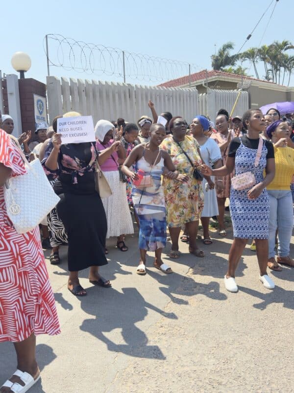 A group of women who are raising white papers with messages against the ill-treatment of the school by the Department of Education. They are also singing songs outside a locked school gate.