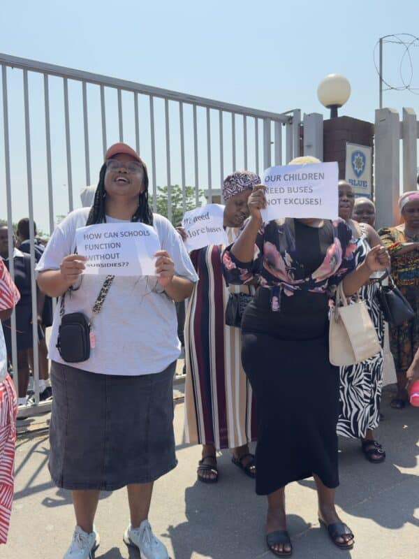 Women protesting outside the gate of a school with signs in front of them and raised. They are also saying something.