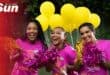 Three women wearing pink t-shirts cheering with pom poms and bright yellow balloons in the background