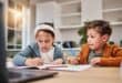 Two children sit at a table at home, writing in workbooks while taking part in an online lesson on a laptop.
