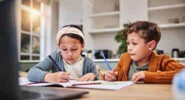 Two children sit at a table at home, writing in workbooks while taking part in an online lesson on a laptop.