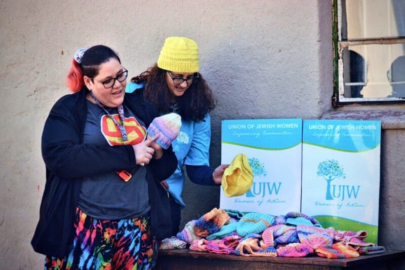 Wheel Well’s Theresa Erasmus shows the children how the halo beanies works at night as Union of Jewish Women’s Vanessa Weltman picks up a yellow beanie from the table in Alexandra on July 17, 2026. Photo: Itumeleng Maloka