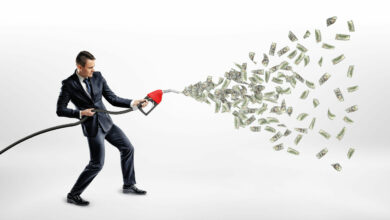 A smiling businessman holding a gas nozzle while many dollar bills are flying out of it instead of petrol.