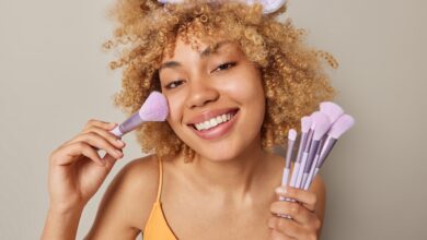 Beautiful smiling woman with curly hair applies blush on face with cosmetic brush powders cheeks looks happily at camera wears t shirt and headband isolated over grey background. Daily makeup