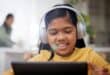 A smiling young girl wearing headphones uses a tablet at home, with an adult blurred in the background.