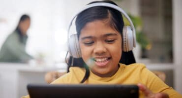 A smiling young girl wearing headphones uses a tablet at home, with an adult blurred in the background.