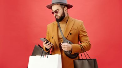 A man is pictured in front of a red back ground holding a cellphone and a few shopping bags.