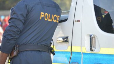 A police officer dressed in his uniform with police written in bold yellow text on his back stands next to a police van.