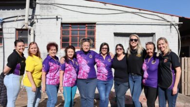 A group of woman dressed in purple pose for a photo. All of them are posed in front of an old, white building.