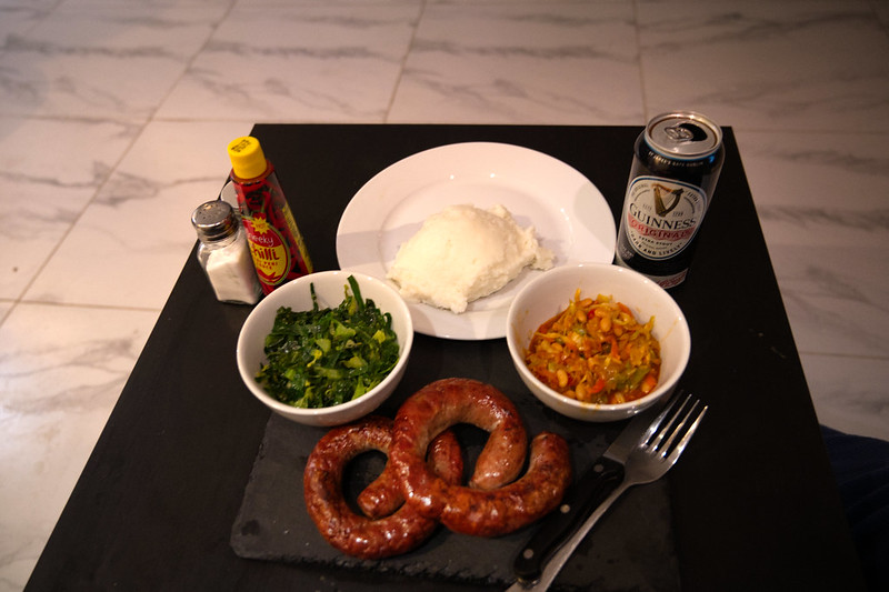 A picture of a meal (Boerewors, pap , chakalaka and garnish) on a table with a can of beer, salt and sauce.