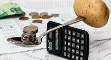 A spoon balanced on a calculator with coins on one side and a potato on the other, symbolising balancing food and money.