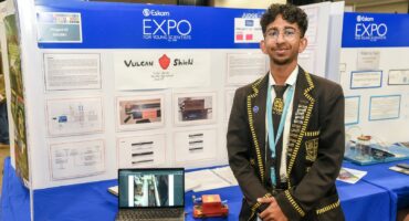 A young man stands proudly in front of a science fair display titled 'Vulcan Shield.' He is wearing a detailed black blazer with gold trim. The booth, part of the Eskom Expo for Young Scientists, includes informational posters and a laptop showing project visuals. A small model vehicle is on the table, enhancing the exhibit's innovative and enthusiastic atmosphere.