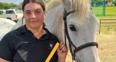 A young white girl, dressed in a black t-shirt holds a yellow medal while standing next to her white horse at a horse-riding rink.