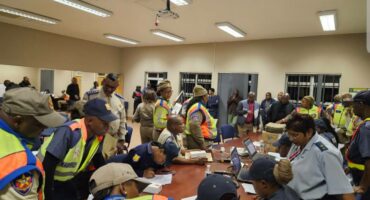 A group of Newcastle traffic officers and SAPS members in bright reflective vests gather around a table in a room, focused on paperwork and laptops after a multidisciplinary operation.