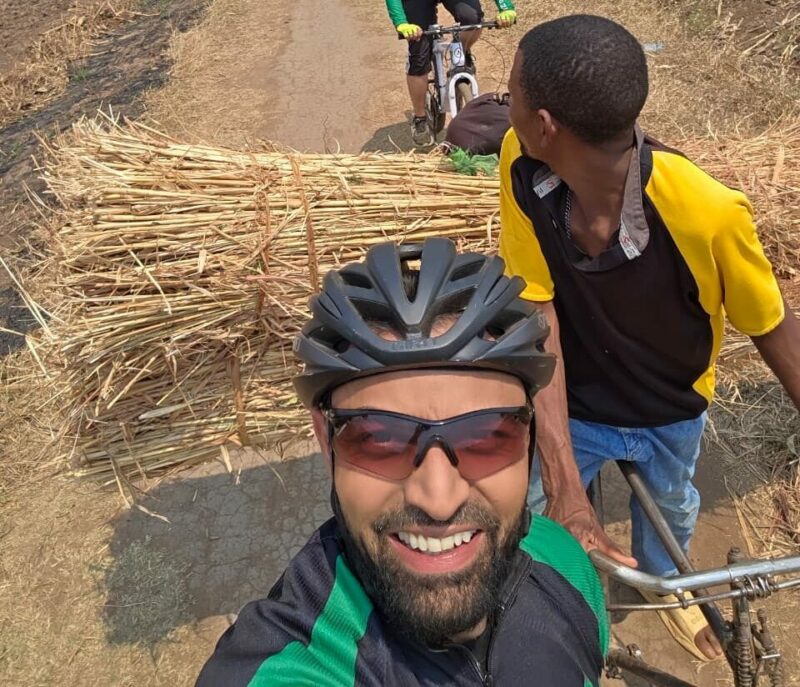 A cyclists travelling on a dirt path in a rural landscape with mountains. A man smiles in the foreground, while a local with a cart full of hay watches. There are two more cyclists pictured in the background.