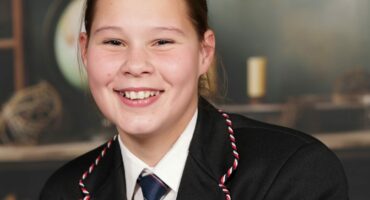 A smiling girl in a school uniform with a striped tie and blazer adorned with pins. Blurred background with books and a globe, creating an academic tone.