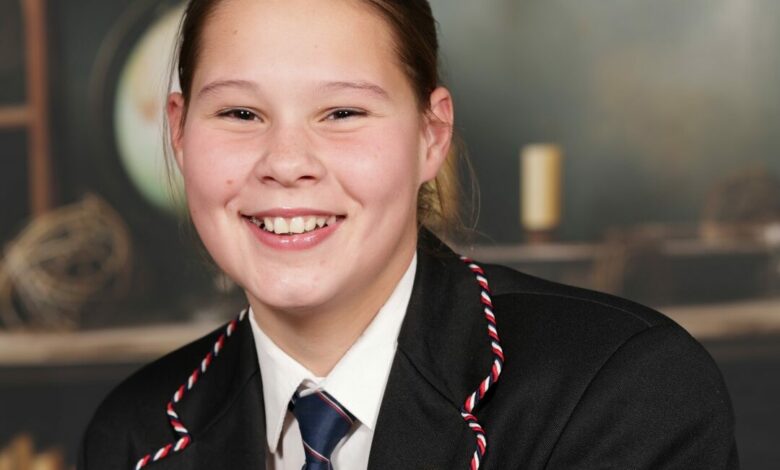 A smiling girl in a school uniform with a striped tie and blazer adorned with pins. Blurred background with books and a globe, creating an academic tone.