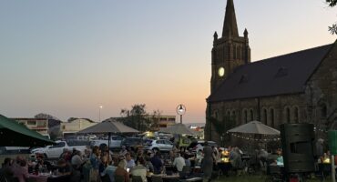 Photo of the NG Klipkerk in the background as the sun is setting with the congregation sitting around tables and fairy lights lighting up the setting.