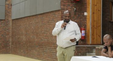 Director Nathi Mbongwa speaks to the community. Photo with black man with white longsleeve shirt and beige pants holds microphone in his hand while speaking to the community.