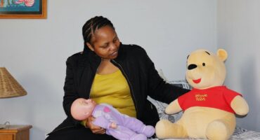 A woman sits on a bed holding a baby doll and looking at a Winnie the Pooh toy beside her.