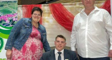 A scholar sits on a couch holding a certificate while his mother and father stand beside him. There is a colourful background with red draping.