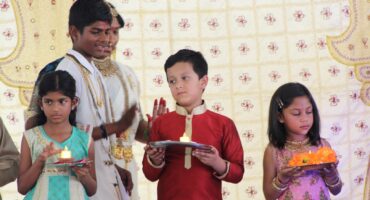 Children dressed in traditional clothing hold plates with lit candles during a cultural ceremony. The background features a decorative patterned cloth.
