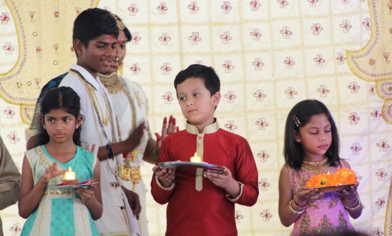 Children dressed in traditional clothing hold plates with lit candles during a cultural ceremony. The background features a decorative patterned cloth.