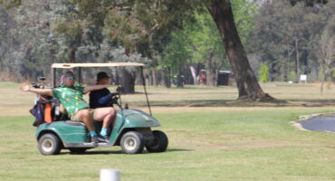 two people riding a golf cart on a green golf coarse with trees seen in the background.