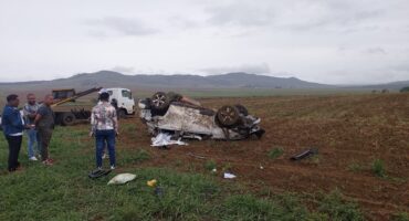A white bakkie on its roof in a field with people standing around looking at it.