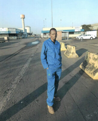 Black man with blue overall standing in the street.