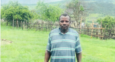 Black man dressed in blue stripe shirt and blue pants standing in a green field.