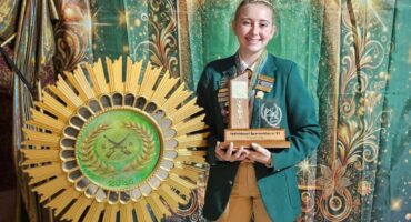 A learner dressed in SA colours with a trophy in her hands and the national target shooting emblem next to her.