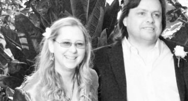 A smiling couple standing together in front of leafy plants, dressed smartly for a celebration.