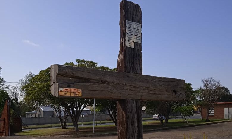 A weathered wooden cross stands in a grassy area, surrounded by plants. The sky is clear and blue, suggesting a calm and serene atmosphere.