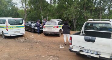 Several police officers stand near marked police vehicles in a wooded area at the Newcastle bus rank, engaging in conversation while at a scene of a deceased person found floating in the river.