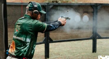 A man is seen firing a pistol at a shooting range. He is wearing a green sports shirt with 'South Africa' text. Gun smoke is also pictured and as a bullet casing is ejected. He is in nature with trees and grass in the background.
