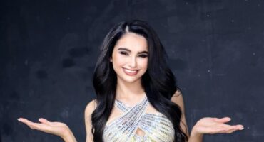 A young girl dressed in a fancy dress standing infront of a dark wall, smiling for the camera.