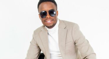 An african man wearing sunglasses and a light beige suit sits on a stool against a white background. He wears a white turtleneck.