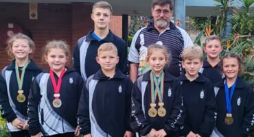 Photo of male and female wrestlers with their medals around their necks with the coach in the back. Dressed in black and white tracksuit jackets.