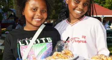 Two young girls smiling for a photo while holding plates of doughnuts. Outdoor background.