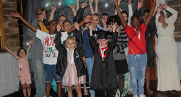 A group of children celebrate academic achievements in front of a black background decorated with colorful baloons.