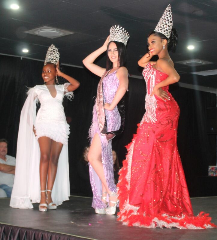 Three beauty queens dressed in beautiful evening wear, wearing sashes and crowns on the Miss Wonder Woman Beauty Pageant Stage.