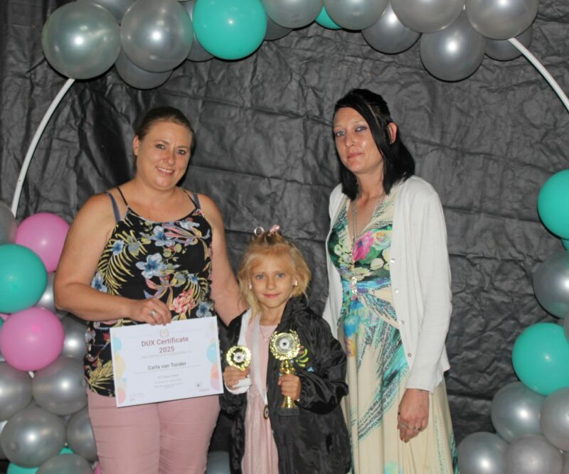 Three girls pose in front of a black and coloured baloon background holding certificates during a prize giving ceremony.