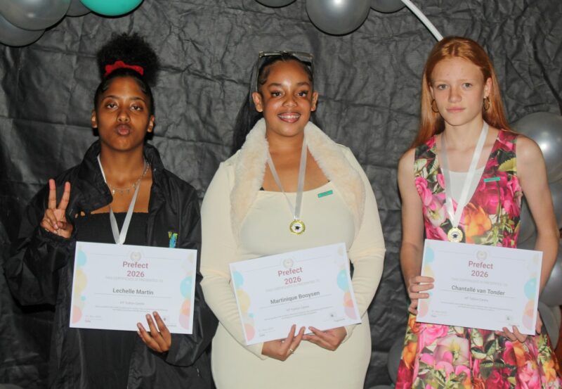 Three girls pose in front of a black and coloured baloon background holding their elected prefect certificates.