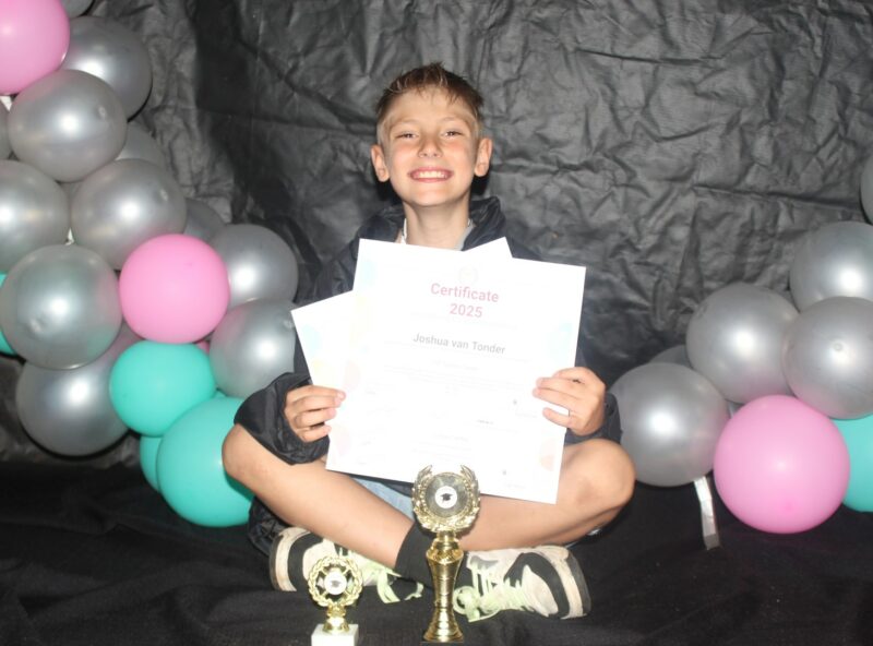 A young boy sits infront of a decorated background holding his certificates with trophies infront of him.