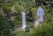 A beautiful photograph capturing the scenic view from Moorfield Farm. Seen in the photos is mountains, blue skies, and green trees and a man is seen standing in the grass with a waterfall in the forground.