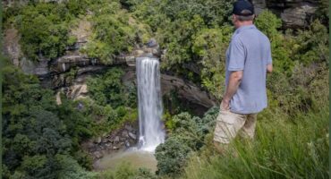 A beautiful photograph capturing the scenic view from Moorfield Farm. Seen in the photos is mountains, blue skies, and green trees and a man is seen standing in the grass with a waterfall in the forground.