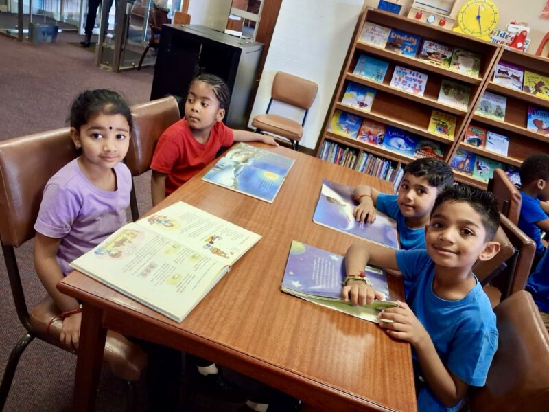 A group of pre primary children dressed in colourful T-shirts sitting at a table in a library.