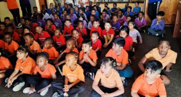 A group of pre primary children dressed in colourful T-shirts sitting on the ground in a library.