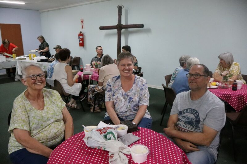 There are three people sitting at a round table. The table has things on it. The table cloth has white and pink squares on it. There is a wooden cross against the wall in the background. There are other people in the background too.